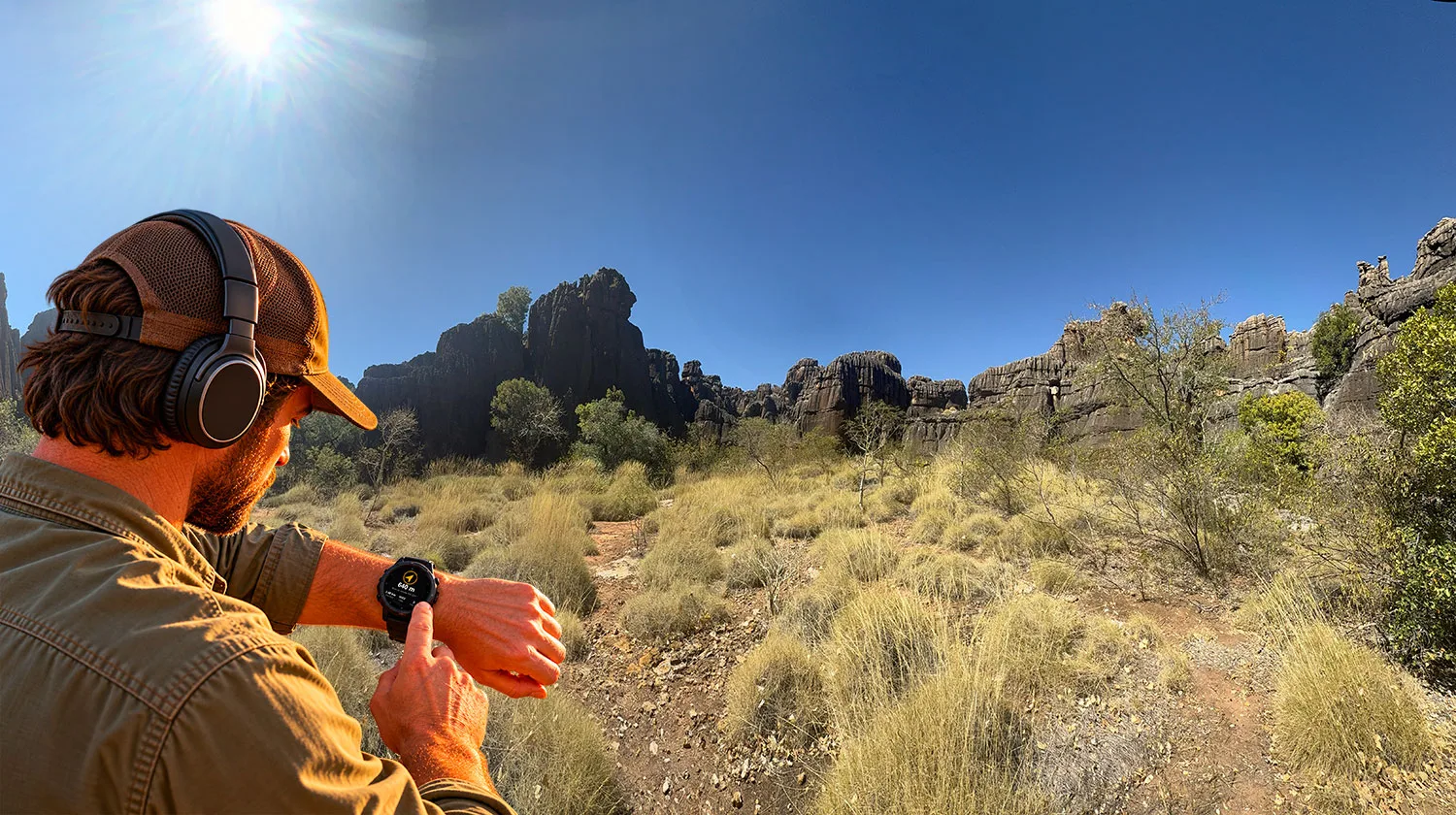 A prospector in a cap and detector headphones checking the way back on his watch, with spinifex and dark rock ranges in Western Australia behind him.